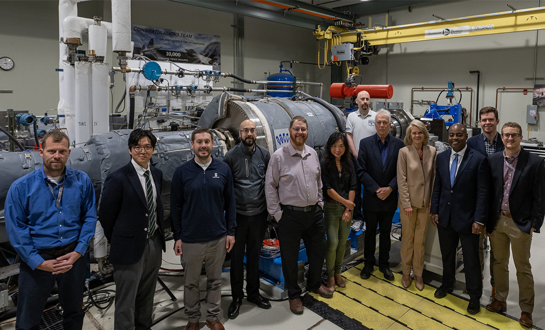 Twelve people stand in front of a turbine in a lab. 