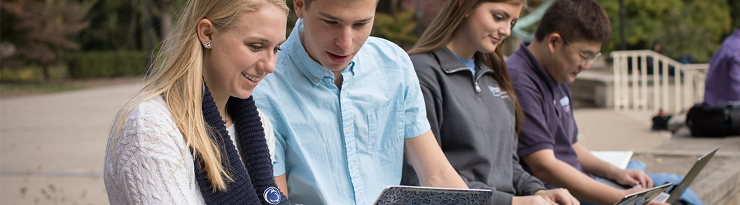 A group of students sit on a bench looking at laptops.